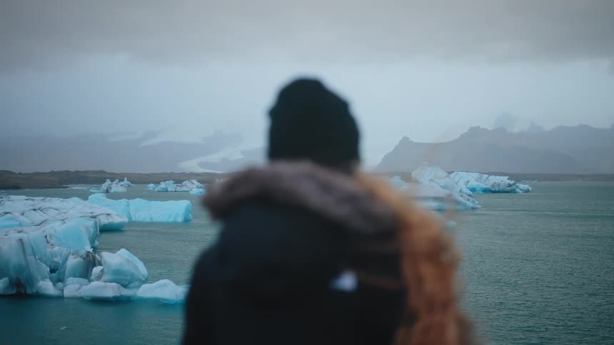 An outoffocus traveler stands before a majestic glacial lagoon gazing out at jagged blue icebergs drifting beneath a moody overcast sky