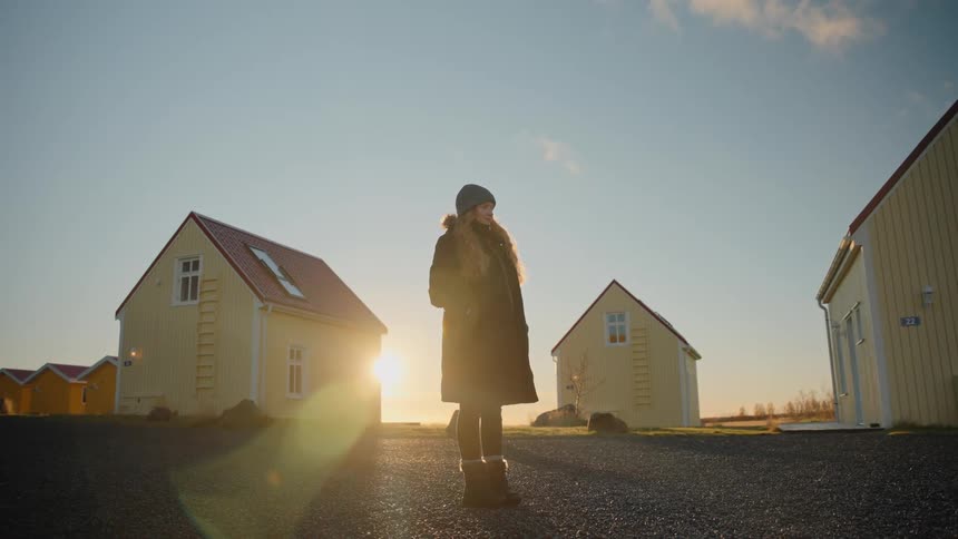 A woman wearing a long winter coat and hat stands outdoors on a gravel path silhouetted by the bright sun setting or rising directly behind her she is framed by a collection of small yellowsided traditionalstyle houses with red roofs in the backgrou