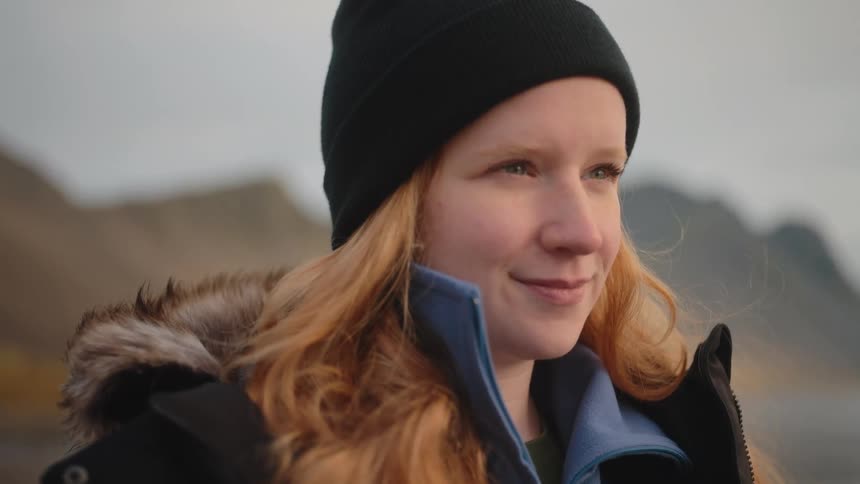 A young woman stands outdoors at midday wearing a winter jacket and a beanie hat smiling warmly