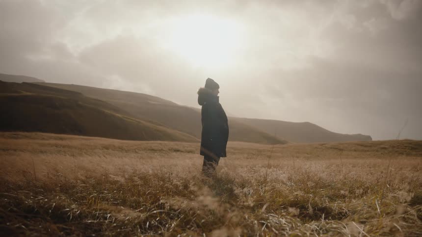 A person stands in a grassy field as the wind blows it around