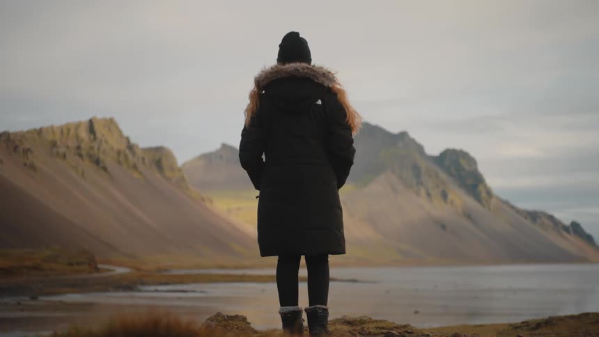 A young woman wearing a winter jacket stands on the bank of a lake at midday gazing across the water as a mountain range lines the background