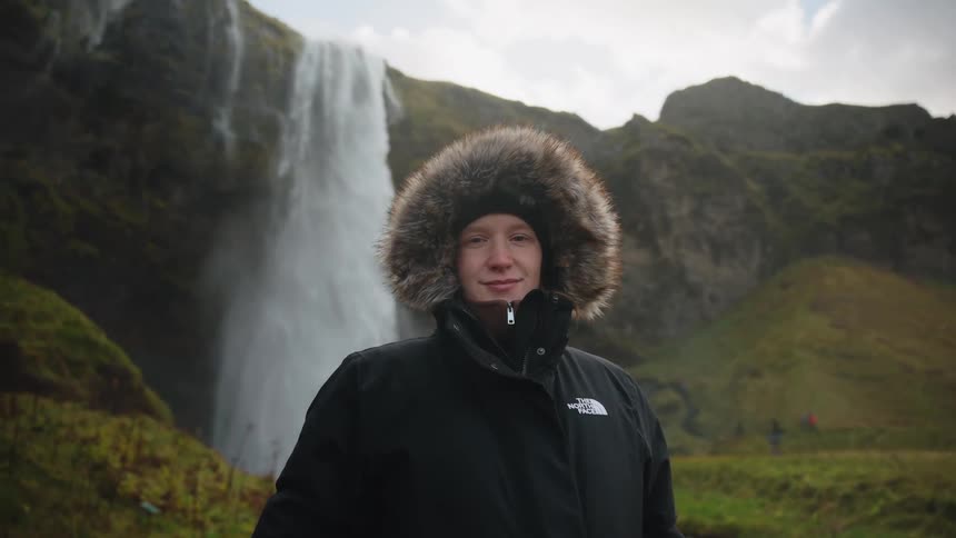 A person wearing a heavy black parka with a furlined hood stands still and looks directly at the camera framed against the backdrop of a large powerful waterfall cascading down a steep mosscovered cliff face