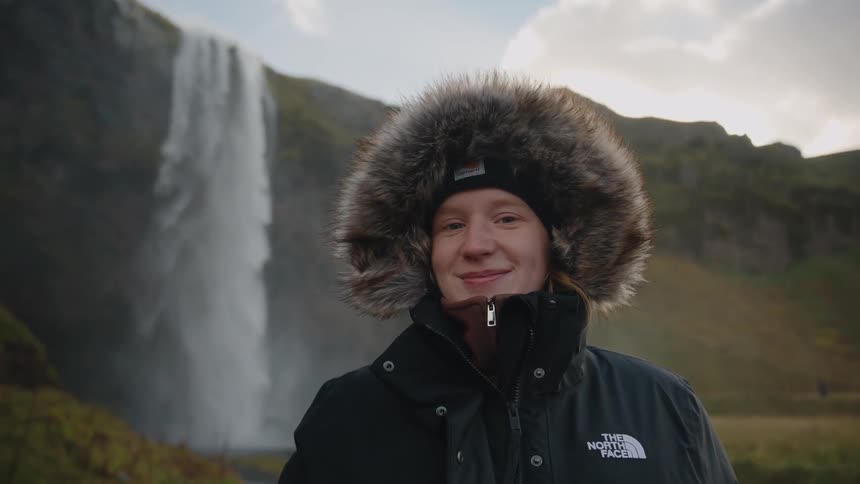 A person wearing a warm winter coat with a furlined hood and a beanie smiles directly at the camera a large impressive waterfall cascades down a cliff face in the background suggesting they are in a cold or mountainous natural environment