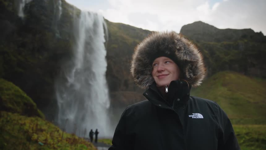 A person wearing a warm black parka with a large fluffy furtrimmed hood is shown in a scenic outdoor setting a powerful waterfall plunges down a rocky cliff face in the background with two small figures walking on a path near the base of the falls