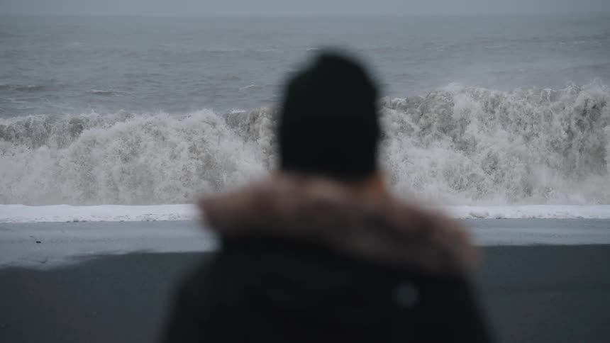 A lone figure in a winter coat stands on a dark beach gazing at the relentless frothing waves