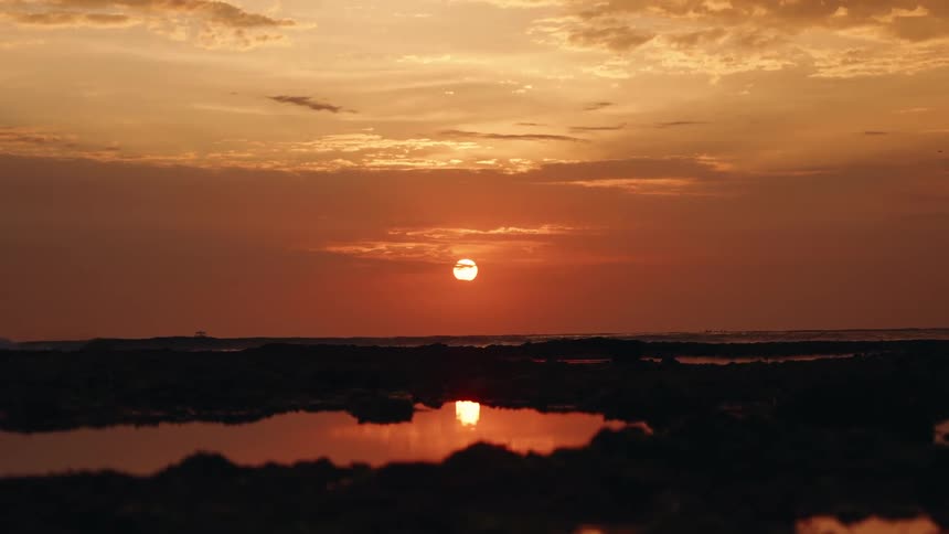 An orange sunset illuminates the sky over a rocky coastline reflecting in the tidal pools and creating a dark silhouette in the foreground