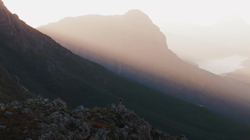 A person is looking forward while standing on a high rocky outcrop against a dramatic sundrenched mountain background showing awe and solitude