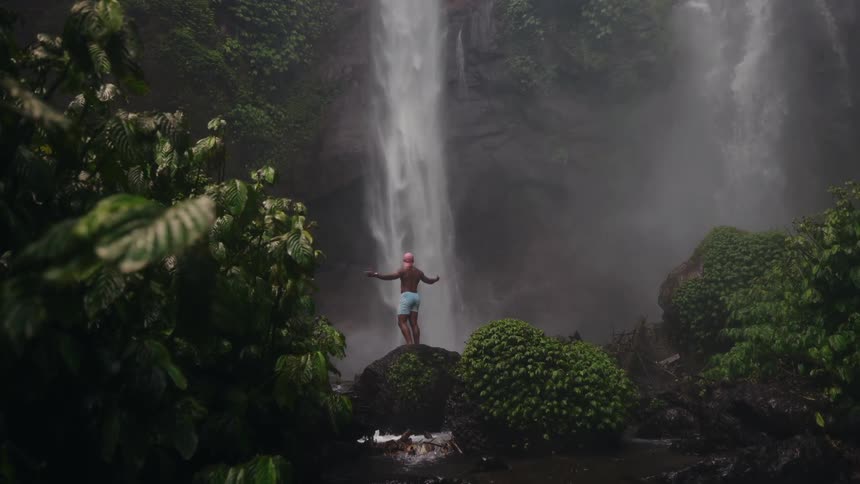 A shirtless man wearing blue shorts stands with arms raised on a large rock in a lush jungle setting facing a tall misty waterfall