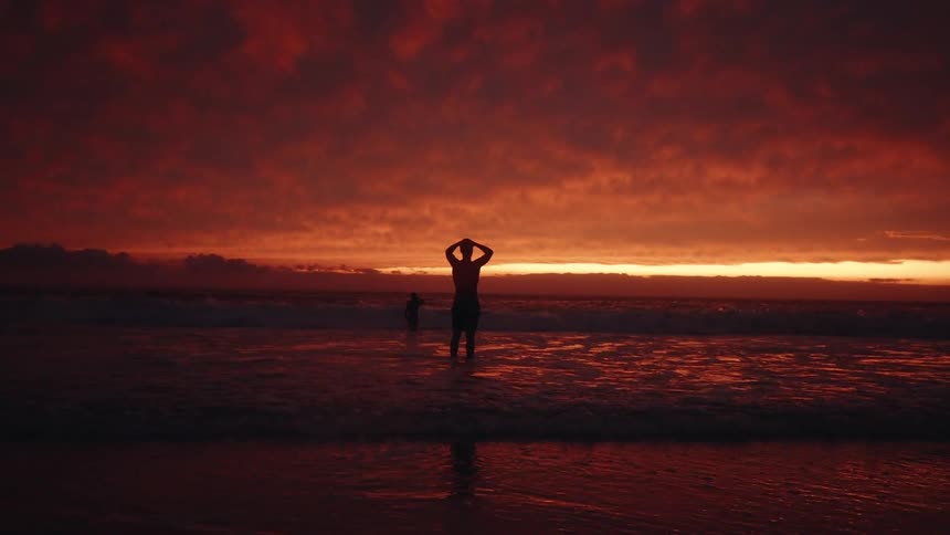 A person stands in the shallow ocean water with their hands raised behind their head silhouetted against an intensely dramatic red and orange sunset sky the water reflects the fiery colors of the clouded horizon creating a warm vibrant tone