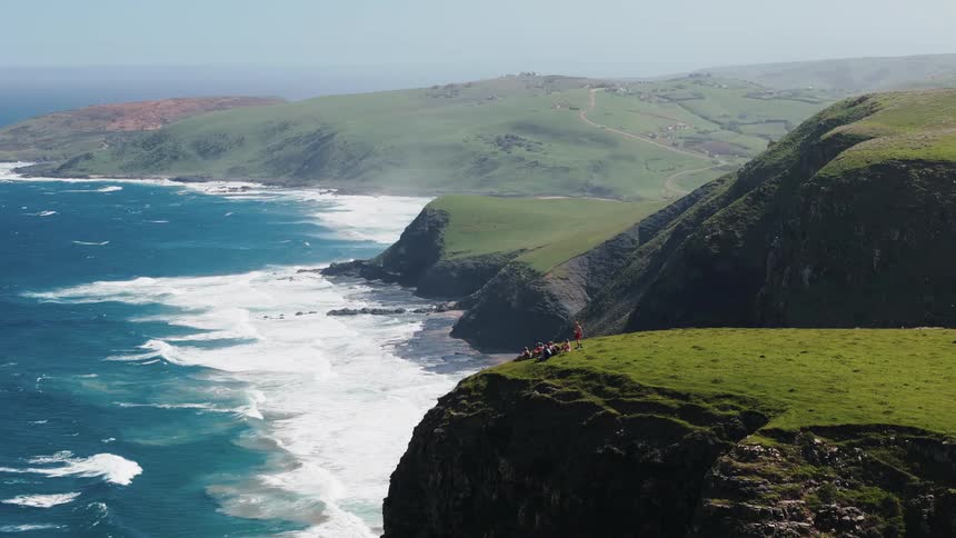An aerial view pans across a dramatic coastline showing steep grasscovered cliffs meeting the turbulent deep blue oceana small group of people sits together on the edge of the vibrant green headland observing the vast coastal landscape and breakin