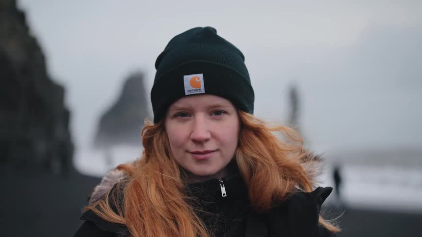 A close view of a young woman with long red hair wearing a black beanie and coat looking at the camera on a dark beach with foggy cliffs in the background