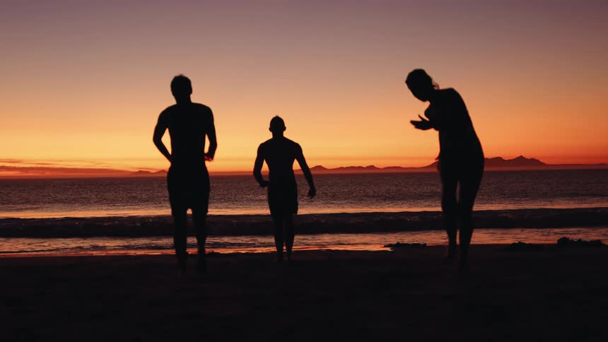 Silhouettes of three young men walking away from the water on the beach under an orange sunset sky
