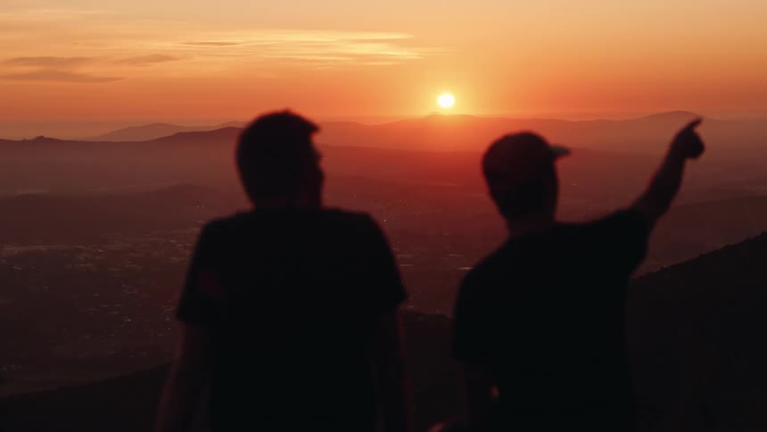 Two men are sitting and looking out at mountains as the sun is low in the sky turning it orange
