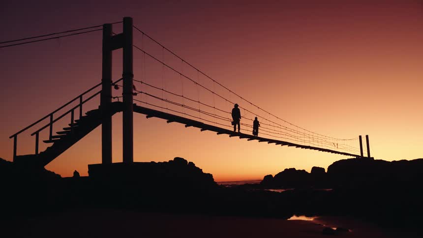 The video is a striking silhouette shot of a long narrow suspension bridge suspended over rocky terrain during sunsettwo figures are visible in silhouette on the bridge slowly walking away from the camera and the access stairs toward the center of t