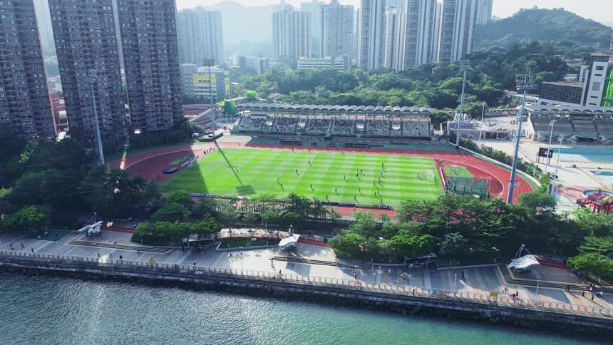This is an aerial view of a stadium with a green sports pitch and a surrounding running track set in a densely populated urban area likely hong kong due to the distinctive tall residential highrises flanking the facility the stadium is located right