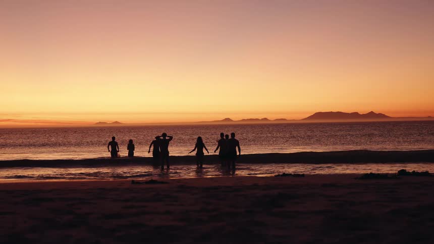 A group of eight people is filmed in silhouette as they walk toward the water and stand at the shoreline on a beach with the ocean and a distant hazy mountain range set against a vibrant orange and yellow sunset sky