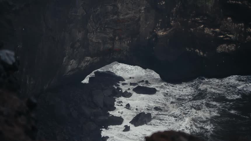 A dramatic highangle shot looks straight down into a dark rocky ocean cove or sea cave opening at the base of a cliff powerful ocean waves surge in crashing against the black rocks and creating thick white sea foam