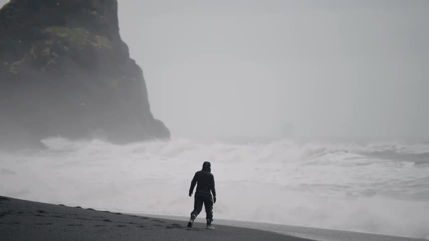 A person wearing a dark jacket and cap walks alone along a vast black sand beach while large dramatic white waves crash against the shore