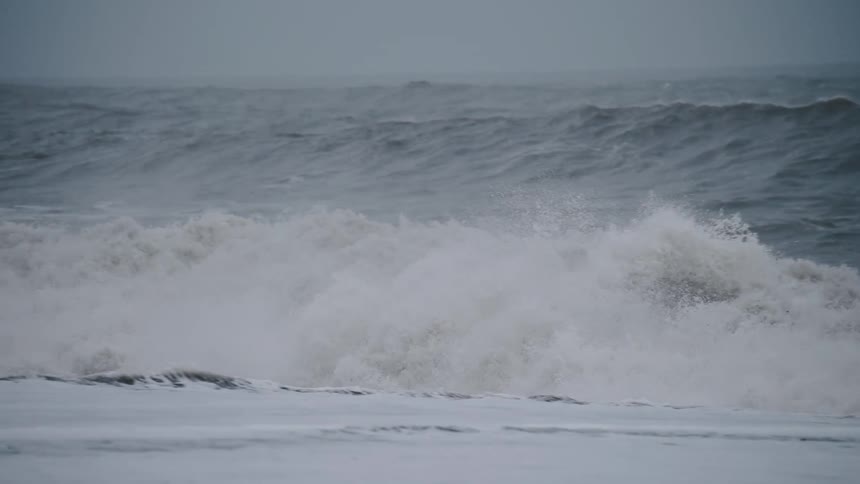 Powerful turbulent ocean waves crash onto the shore creating a massive wall of white foam and spray the scene is dominated by a monochrome palette of greys and whites suggesting stormy weather or a cold overcast day