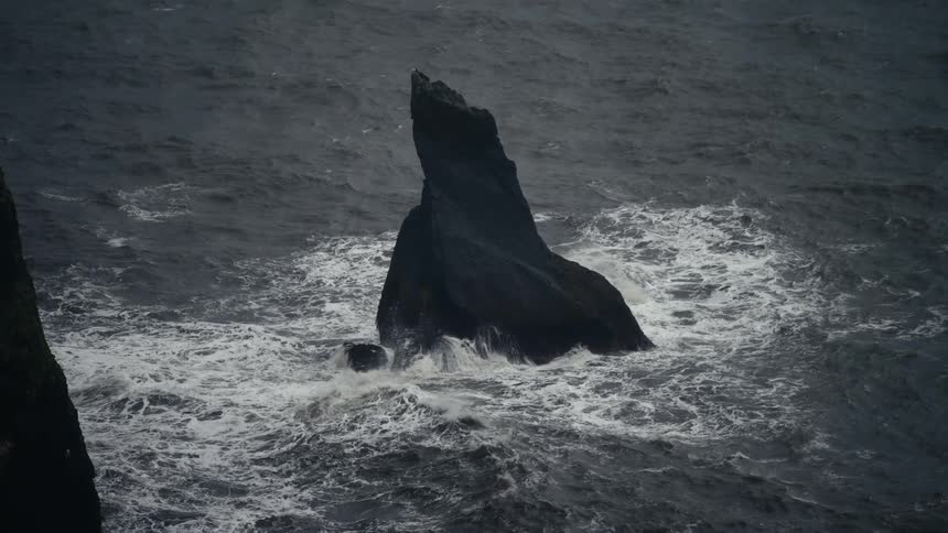 A dark jagged sea stack rises from the ocean with crashing waves at its base