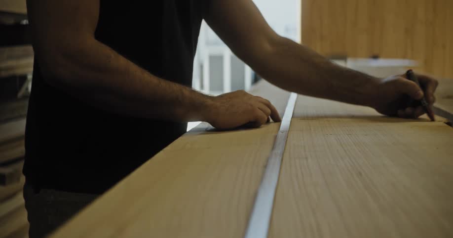 A woodworker is shown marking measurements on a long lightcolored wooden plank using a ruler and a red pencil the initial closeup shows precise marking which then transitions to a wider view of the person working on the large piece of lumber in a wor