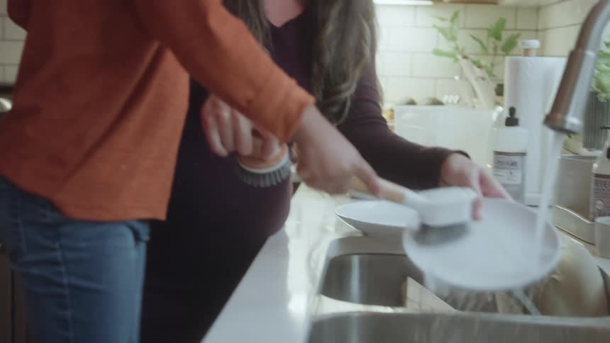 The video shows a warm moment between a woman and a young boy likely in a kitchen where they are washing dishes together at a sink the boy wearing an orange shirt is holding a spatula and assisting the woman who is smiling cheerfully at him