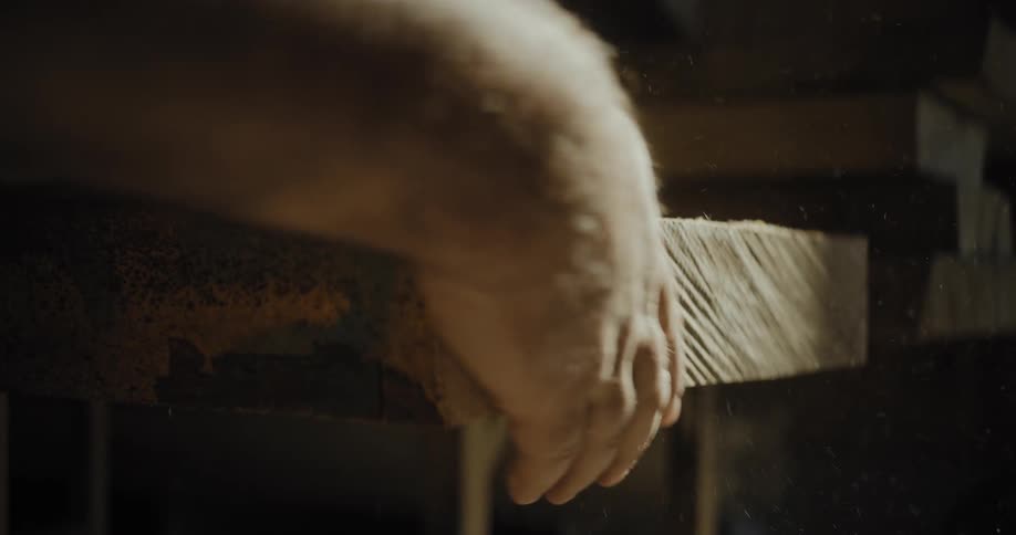 A close view shows a carpenters hand cleaning a thick piece of raw cut lumber with wood dust illuminated against a dark background