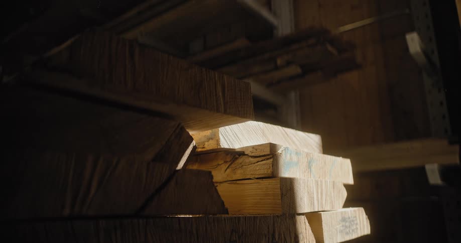 A camera focuses on large stacks of lightcolored raw timber planks stored on wooden shelving in a dark warehouse or lumberyard dramatically illuminated by bright natural light slicing through the darkness