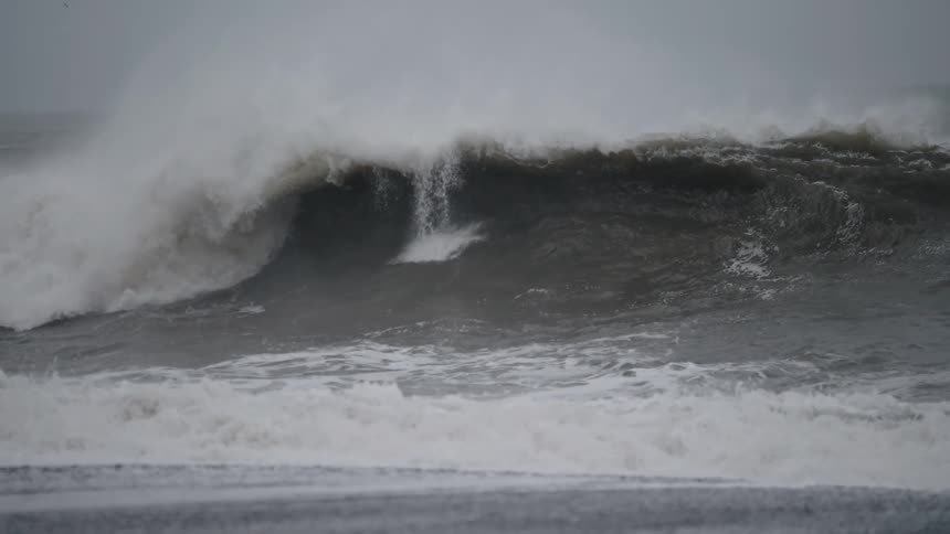 A large powerful ocean wave with dark turbulent water curls and crashes toward the viewer producing significant white spray and seafoam against a dreary overcast sky in a demonstration of rough sea conditions