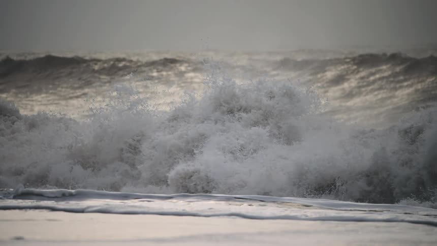 Large dramatic waves crash heavily onto the dark sandy shore under a gray sky creating thick white foam along the beach