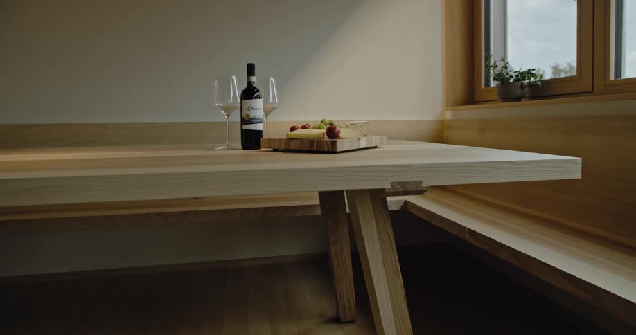 A bottle of wine and a cheese board sit on a wooden table near a window
