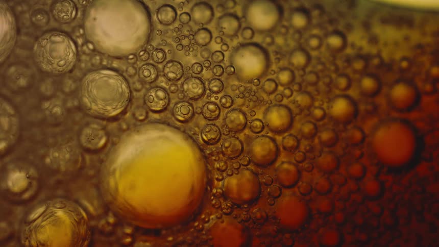 An extreme close abstract view of various sized dark and amber bubbles floating and moving across a rich brown and yellow liquid surface