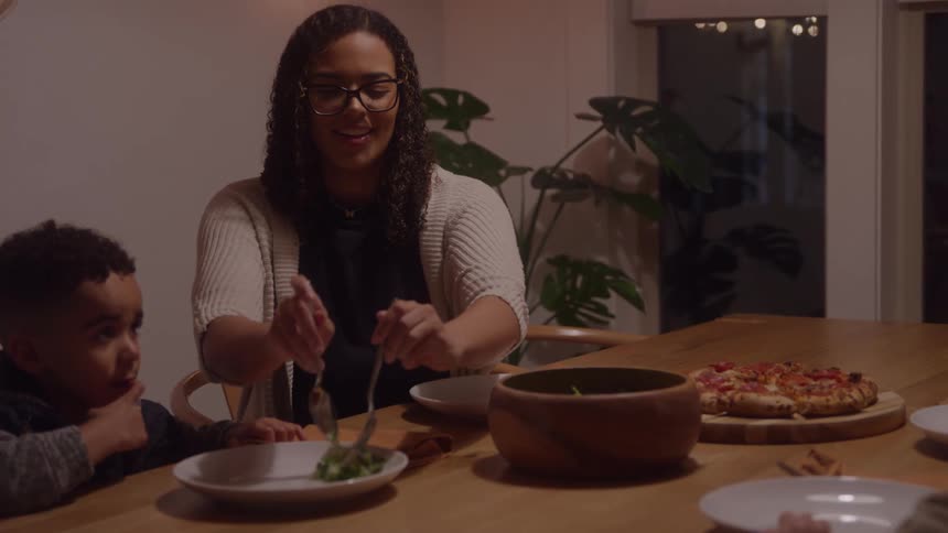 A mother wearing glasses serves salad into her young sons plate during a dimly lit family dinner at a wooden dining table