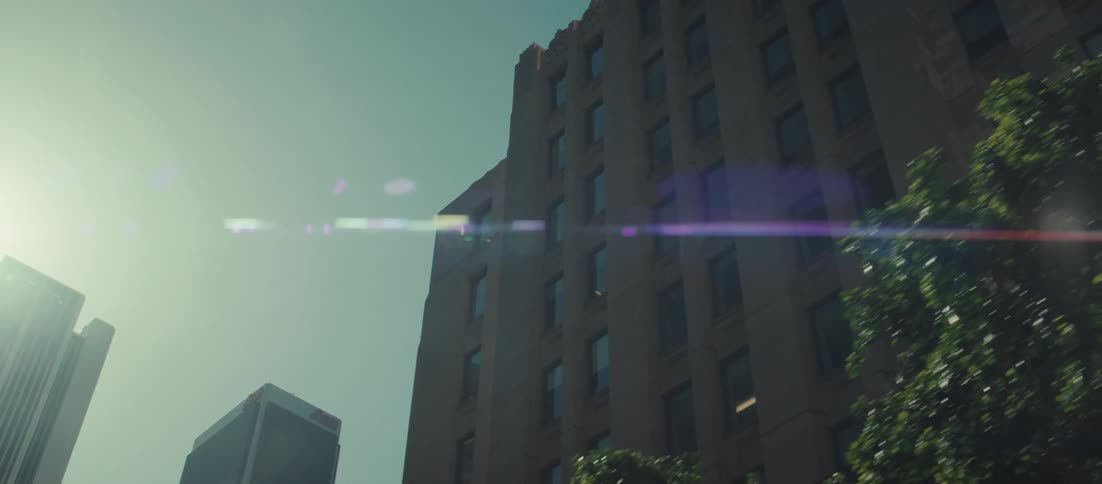 A lowangle tracking shot looks up at towering skyscrapers against a bright sky featuring a prominent lens flare that streaks across the frame