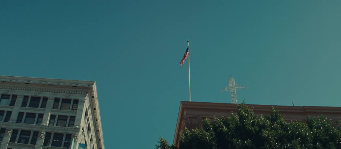 An upwardtilted shot captures two historic city buildings with an american flag atop the shorter structure as the flag is initially flapping gently against a bright clear blue sky
