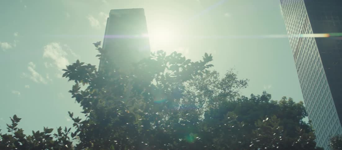 A low angle tracking shot moves through the city showing a tall dark skyscraper silhouetted by bright sunlight and dramatic lens flares against a cloudy sky