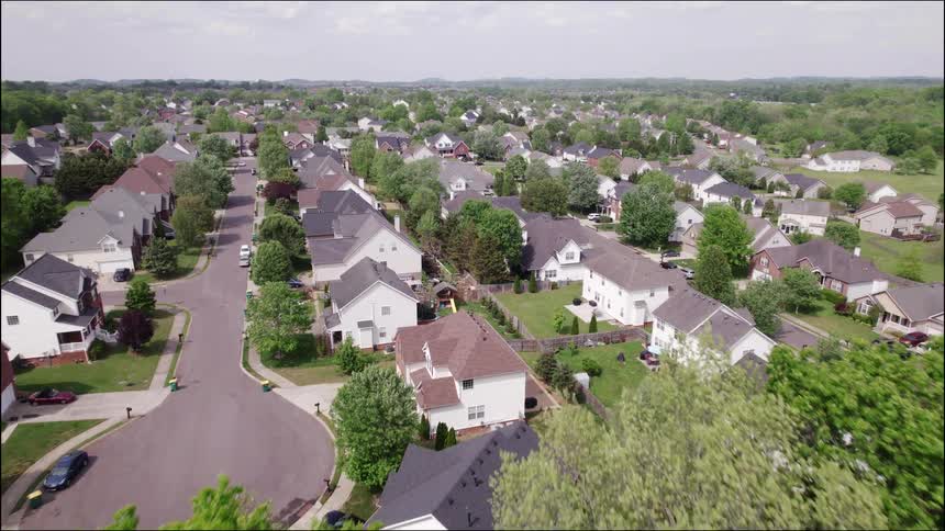 An aerial drone shot slowly descends and moves forward over a quiet wellestablished suburban neighborhood the area is densely packed with houses featuring lightcolored siding and dark roofs interspersed with numerous mature green trees