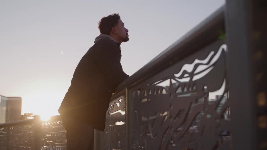A pensive man leans against a bridge railing at sunset slowly lifting his head to gaze at the city skyline