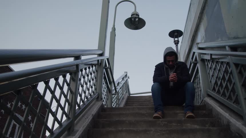 A person wearing a dark hooded jacket and jeans is sitting alone on a concrete staircase outdoors they appear distressed or contemplative holding their hands together and then briefly covering their face