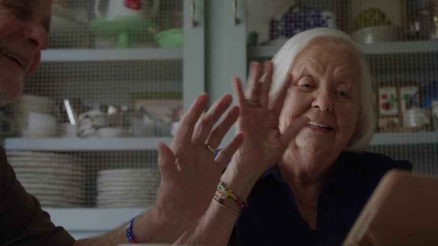 An elderly woman with white hair is seated next to an elderly man gesturing with her hands close to her face in an expression of joy and surprise while sitting in a kitchen with light blue cabinets filled with dishware behind them