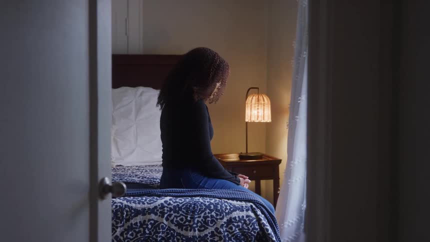 This video shows a young woman sitting quietly on the edge of her bed seen through an open doorway with her back mostly to the camerashe appears contemplative in the softly lit room where a unique caged lamp illuminates the nightstand beside her