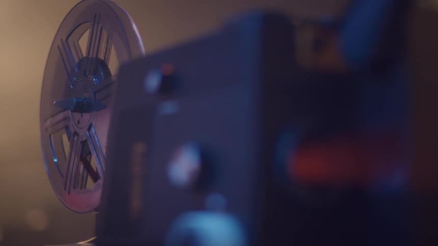 A closeup shot focuses on the large takeup reel of a vintage film projector which is slowly turning in a dark or dimly lit room the projectors internal mechanisms are out of focus in the foreground with warm moody light illuminating the reel