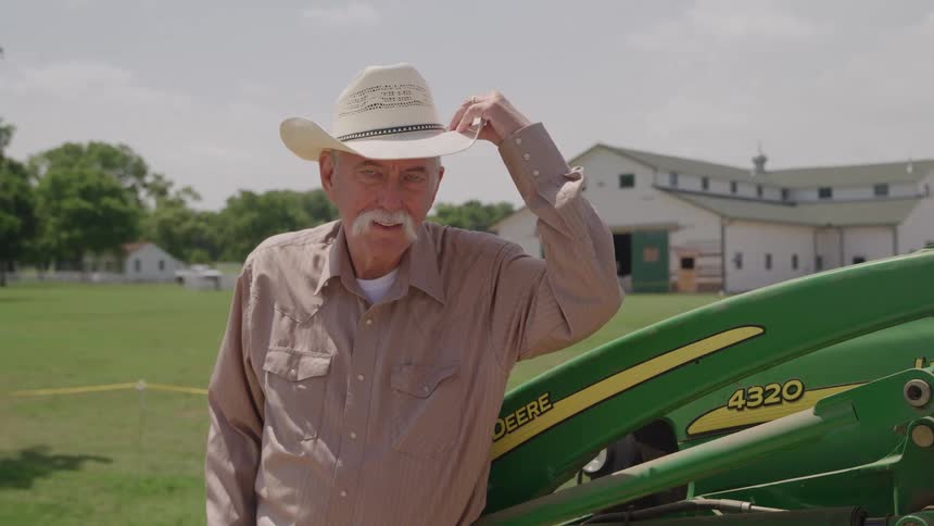 A senior man likely a farmer or rancher stands in front of a barn or large farm building on a sunny day he is wearing a buttonup shirt and blue jeans has a white mustache and places a straw cowboy hat on his head while leaning on a piece of john dee