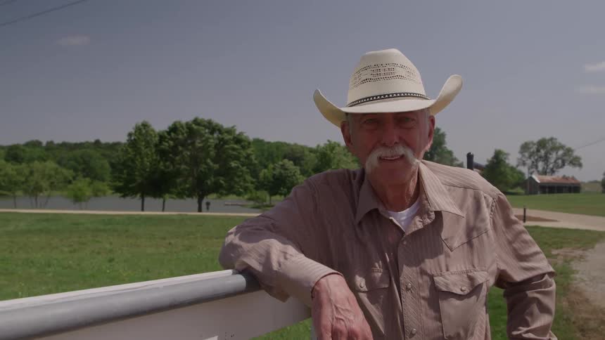 An older man in a cowboy hat leans on a white fence in a rural landscape