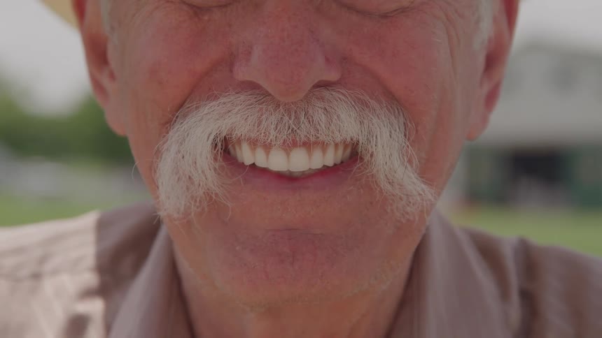 A caucasian male with a large white mustache looks forward with a smiling emotion showing white teeth outdoors near a building