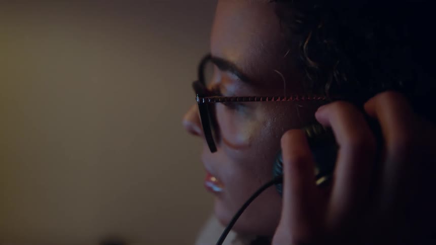 A young woman sits inside an office wearing glasses as she operates a computer and puts an earphone in her ear to listen to sound