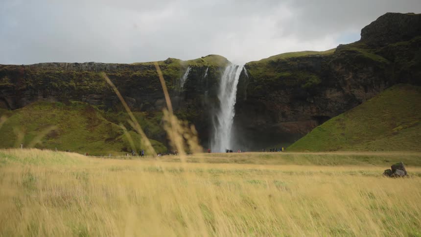 Tall waterfall flows down mossy cliff facing grassy field with people in the distance