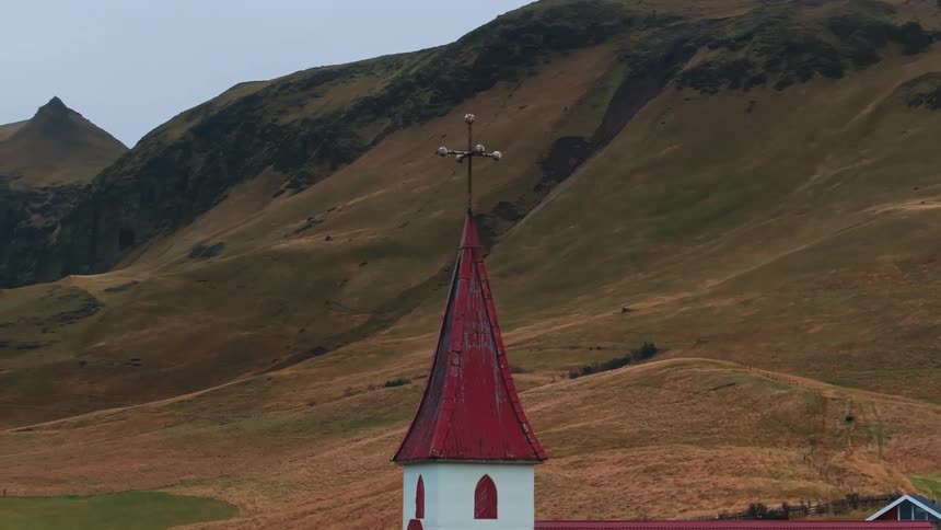 The top of a traditional white church with a tall pointed red roof and a cross on its spire situated in a dramatic landscape of steep brown and green grassy hillsides that dominate the background