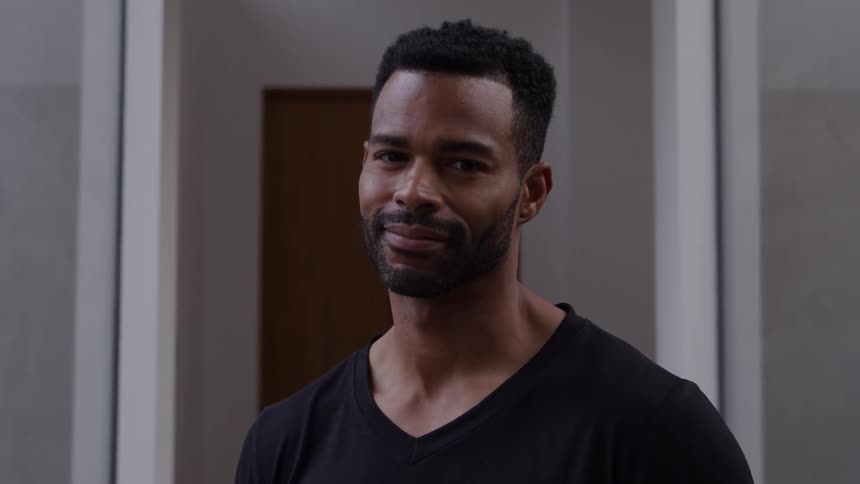 A black male is standing facing forward and smiling in a hallway
