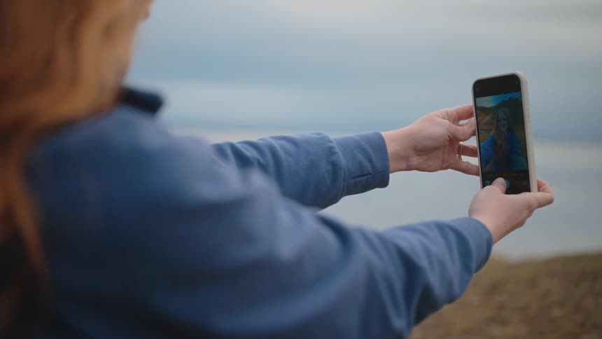 Woman holds a phone taking a selfie in an open area near the sea